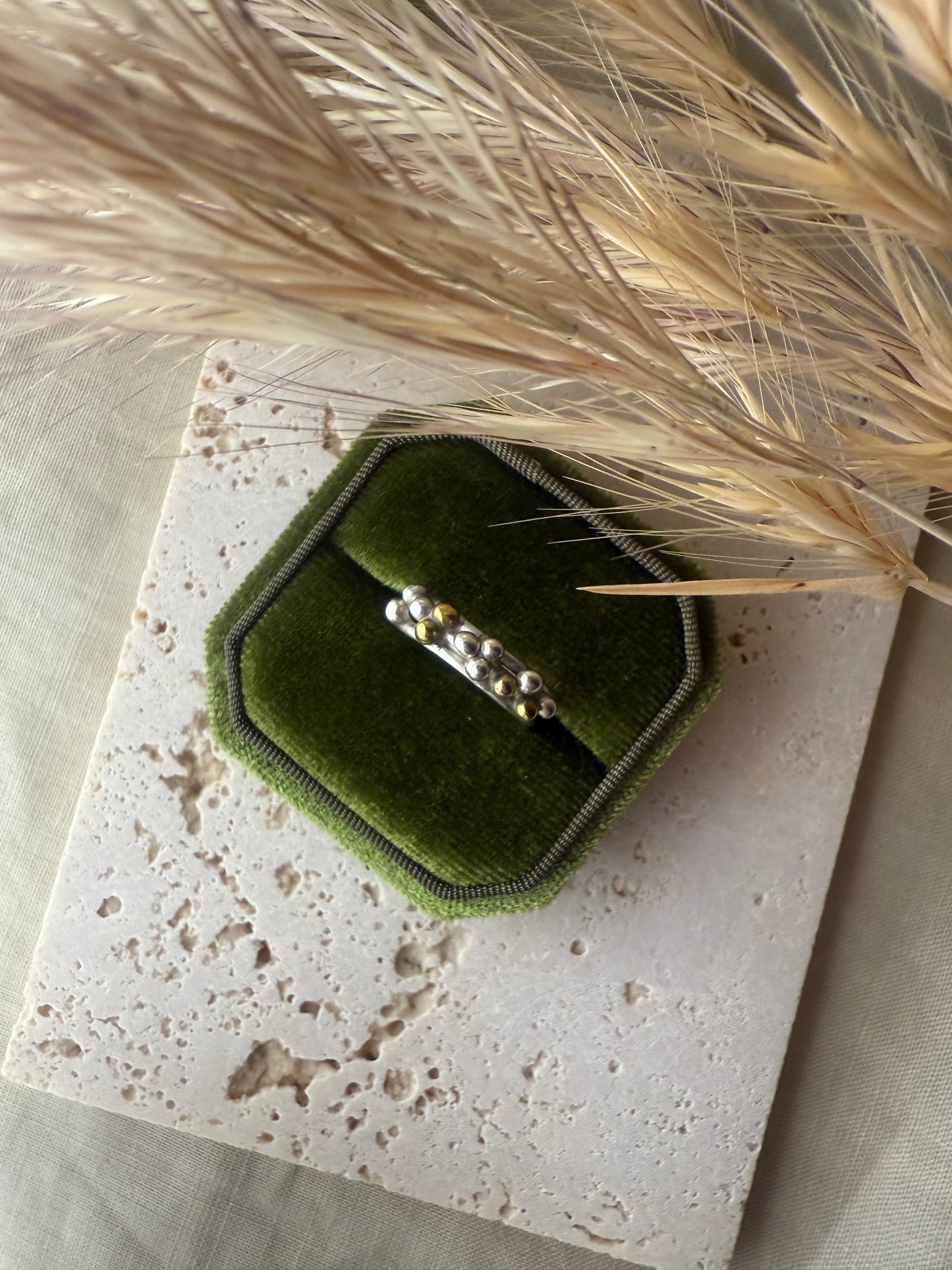 Green velvet ring box with a ring on a textured surface with dried grass.